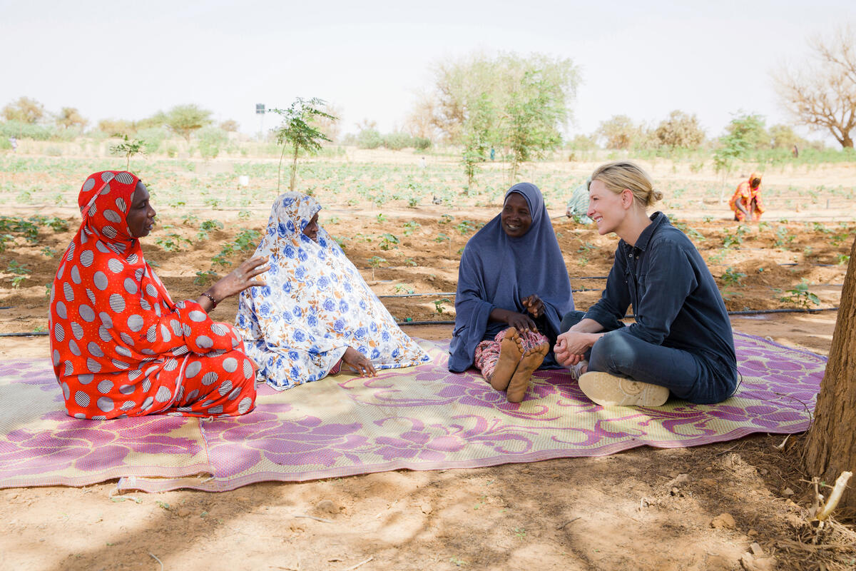 Four women laugh together sitting on a blanket in a vegetable garden.