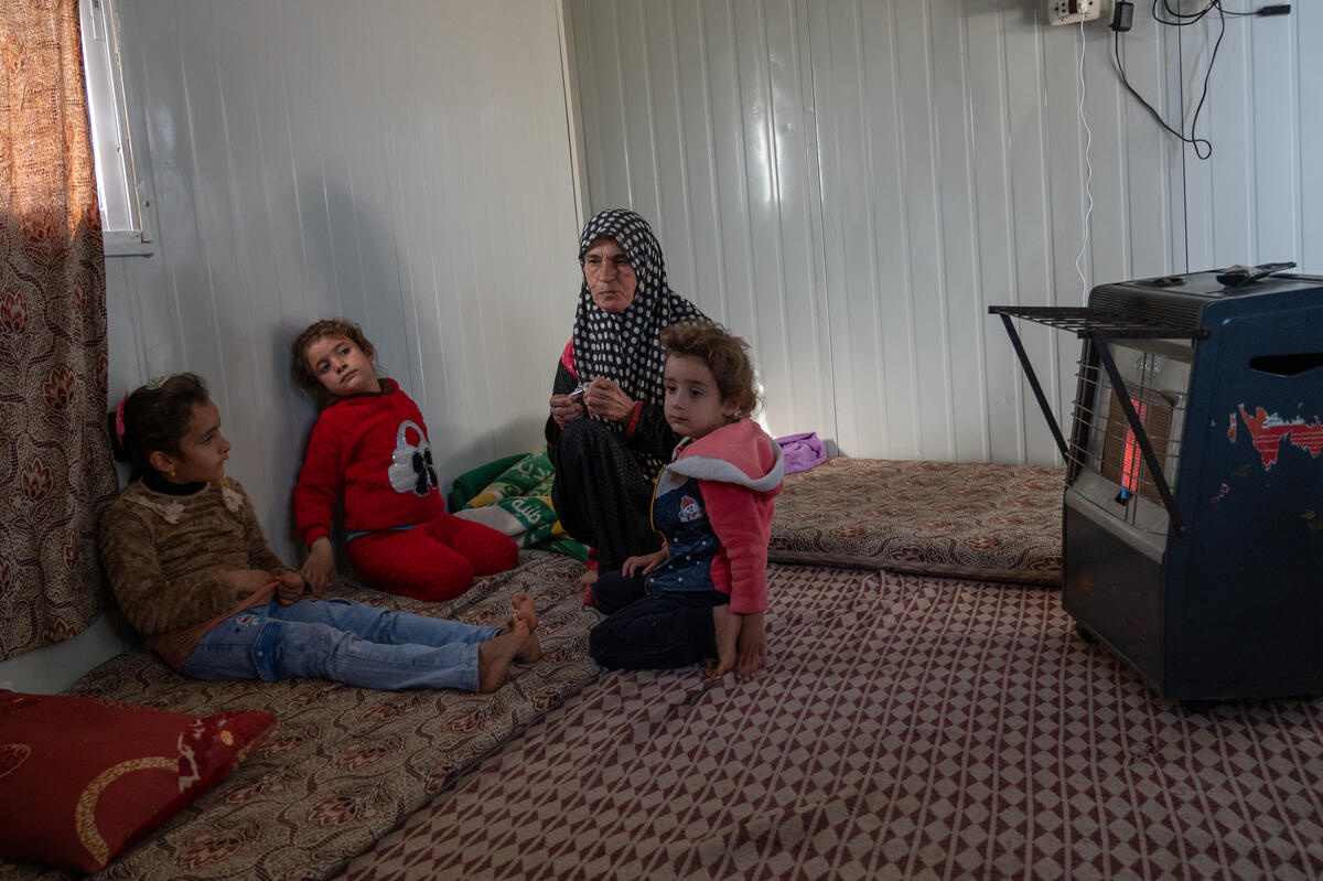 A woman sits with her grandchildren in front of a gas heater inside her shelter.