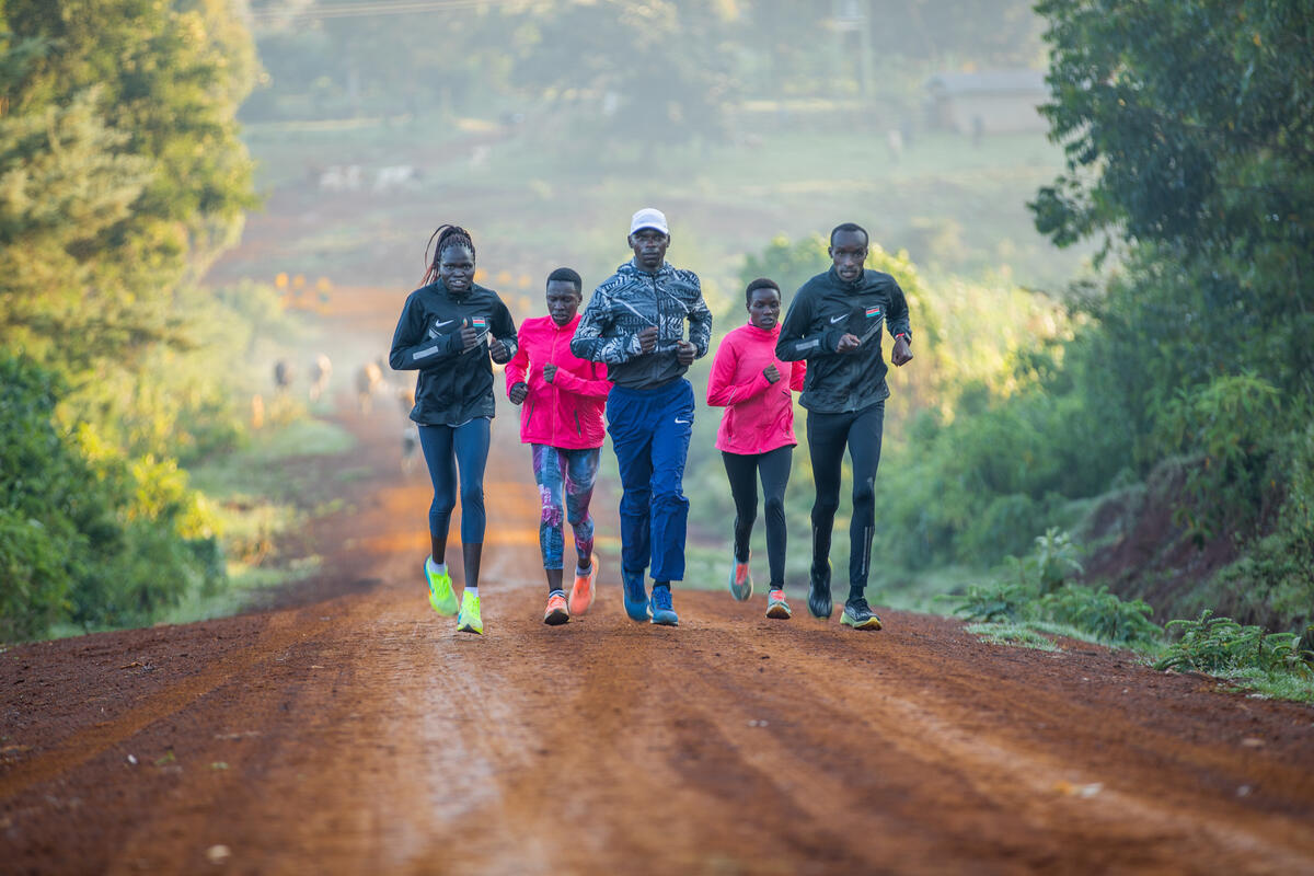 A group of athletes run together down a dirt road in a rural area.