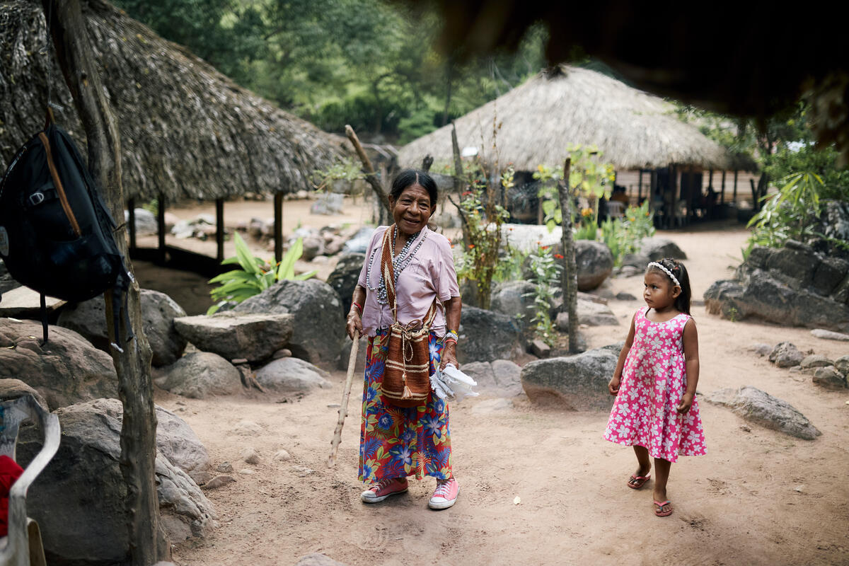 Une femme âgée de petite taille, munie d'un bâton en bois et d'un sac tressé, se tient dans une petite clairière à côté d'abris traditionnels au toit de chaume, sous le regard d'une jeune fille.