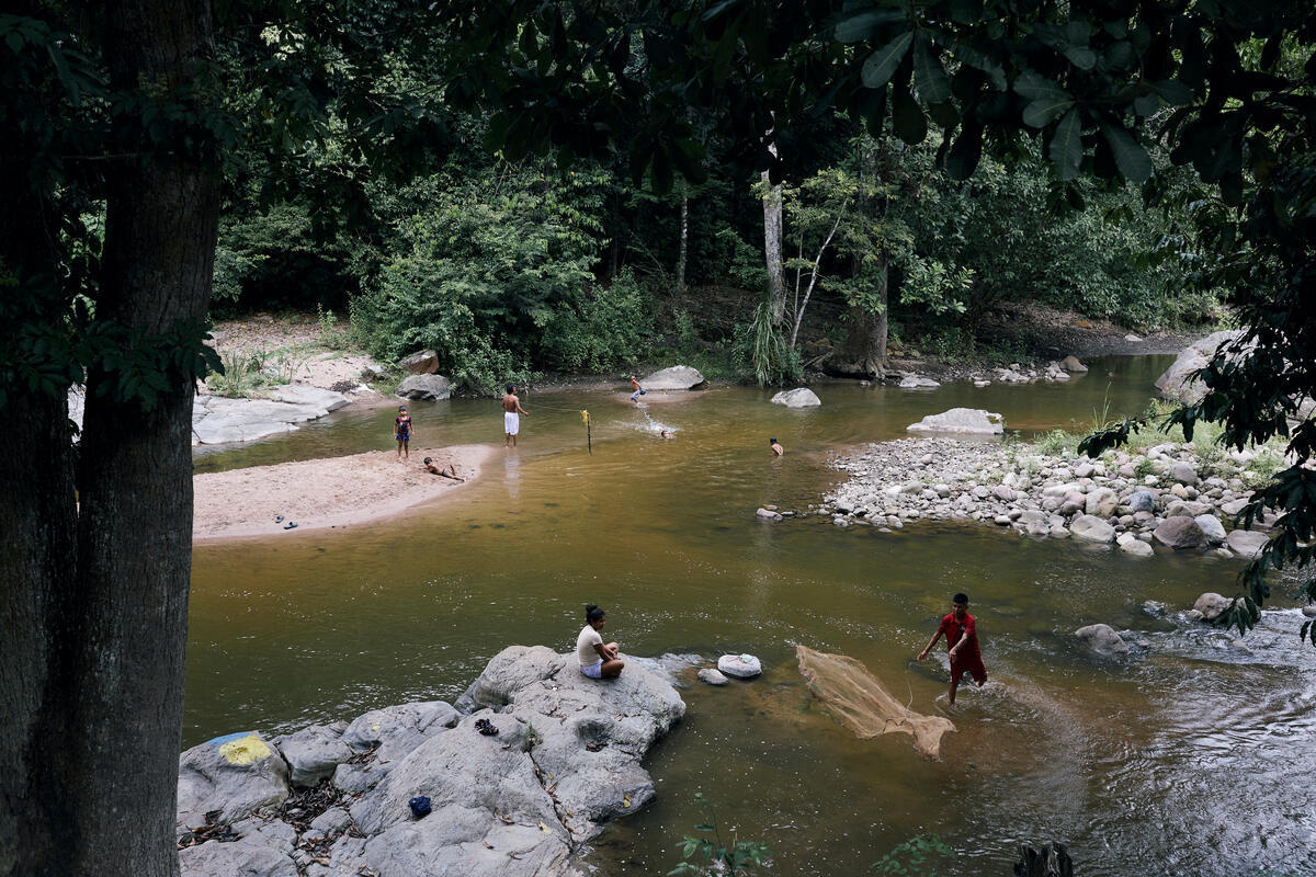 Une vue aérienne d'une rivière montre un homme jetant un filet de pêche et des enfants nageant à proximité, tandis que d'autres personnes regardent depuis les berges.