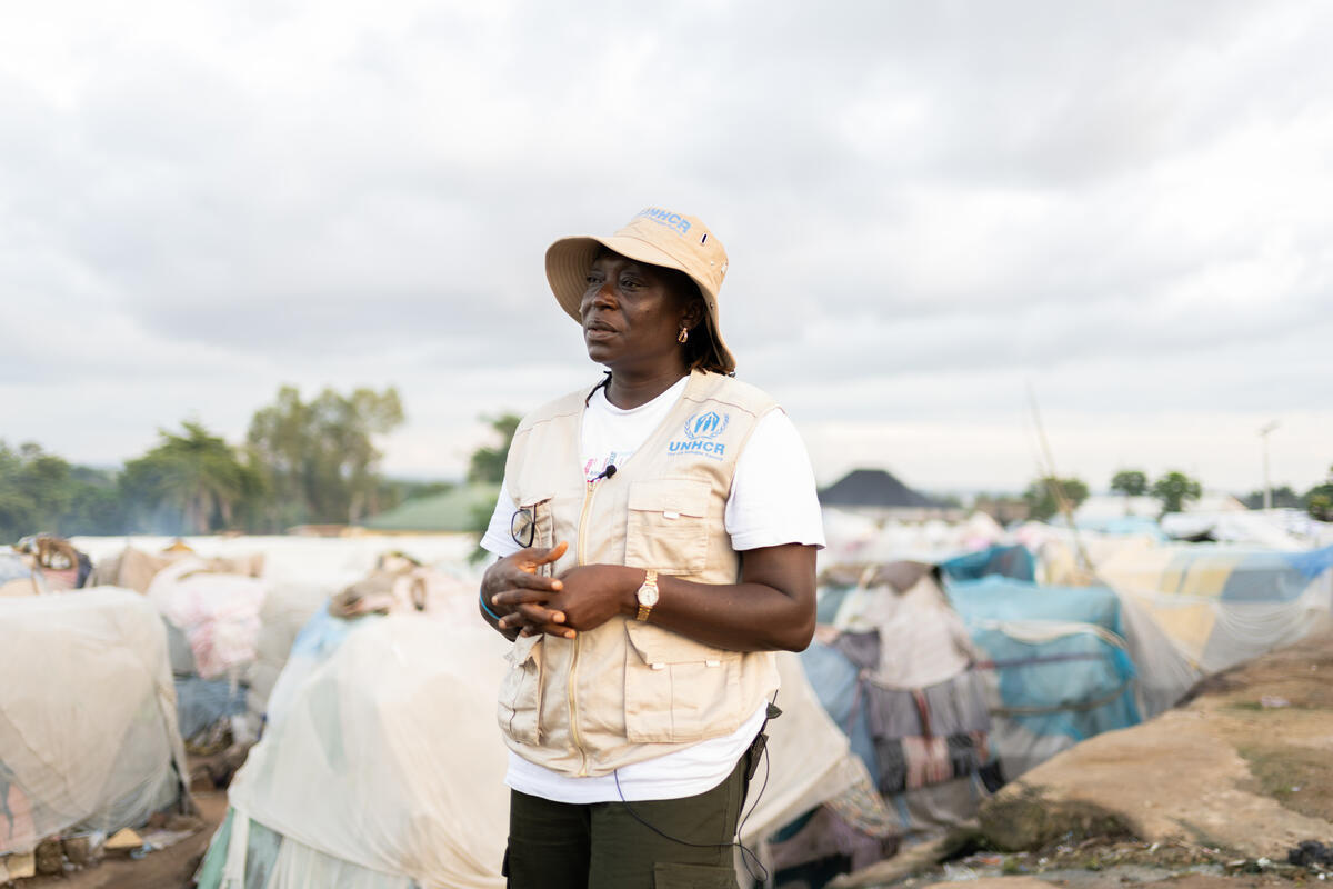 A woman wearing a UNHCR-branded hat and vest stands in a camp for displaced people.