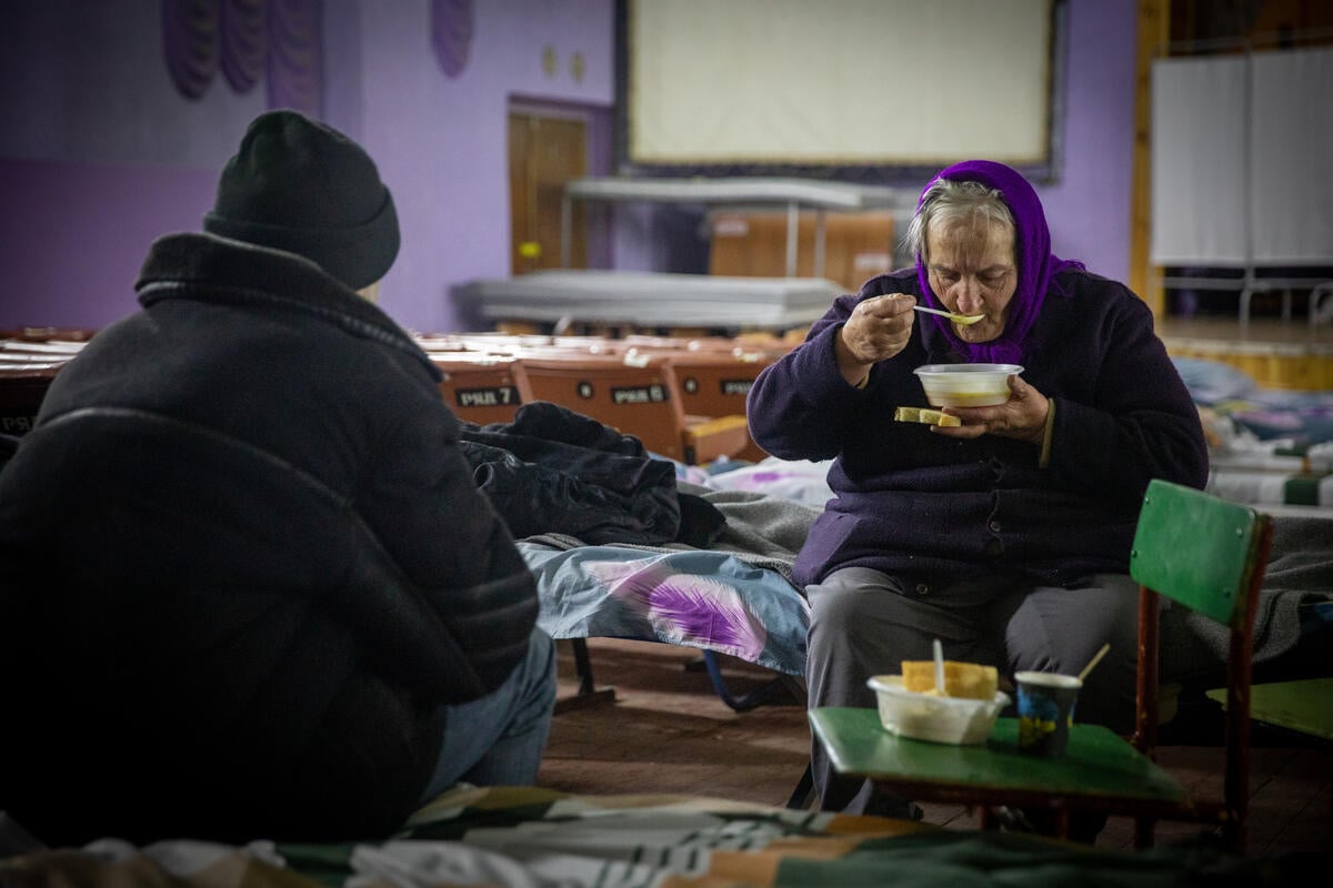 Une femme âgée est assise sur un lit de camp et mange de la soupe et du pain.