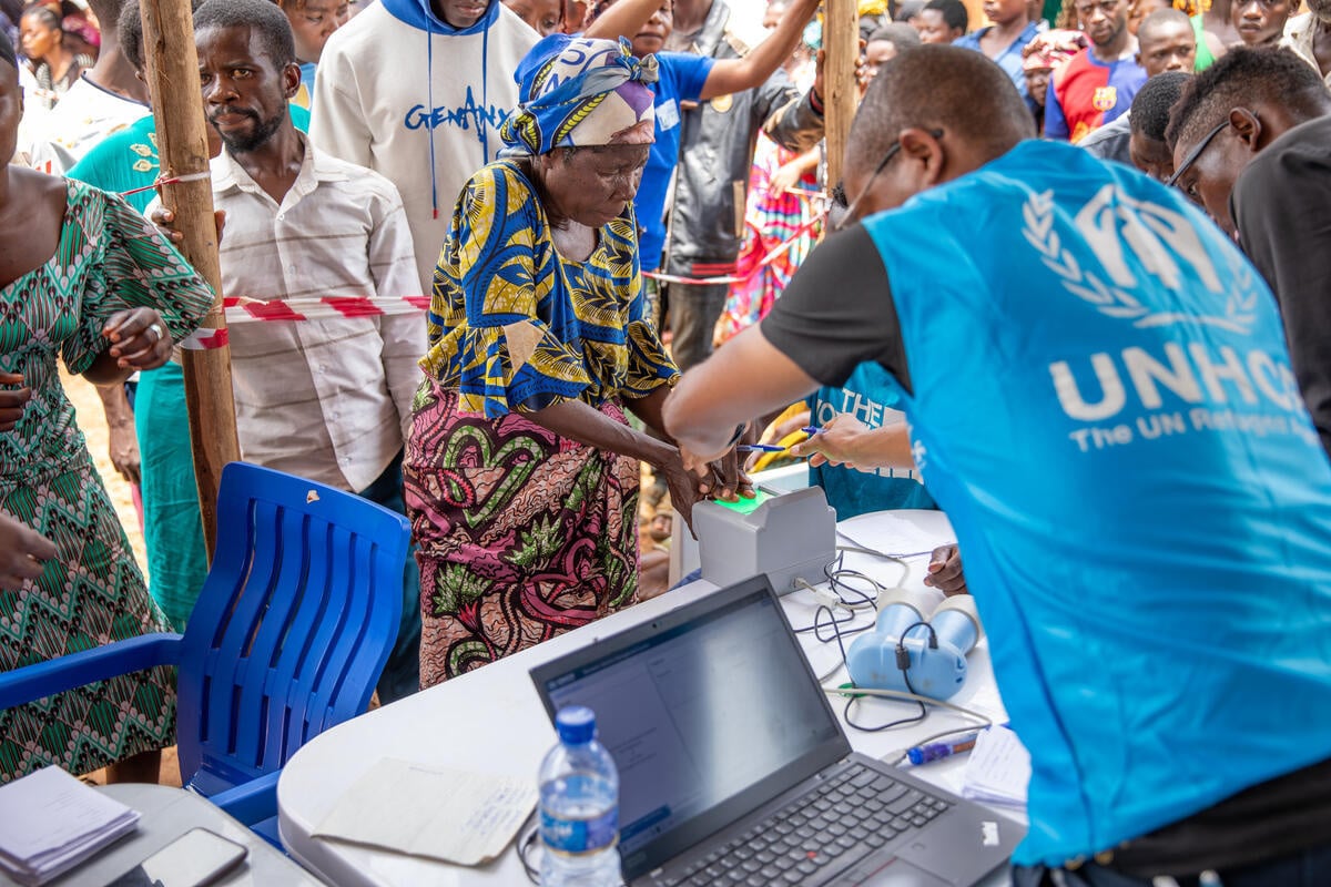 A UNHCR staff member scans an elderly lady's fingerprints at a registration desk while other refugees wait their turn.