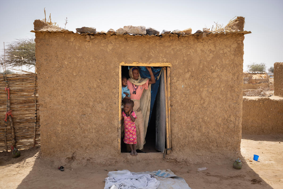 A mother stands with two children in the open doorway of a small mud brick house