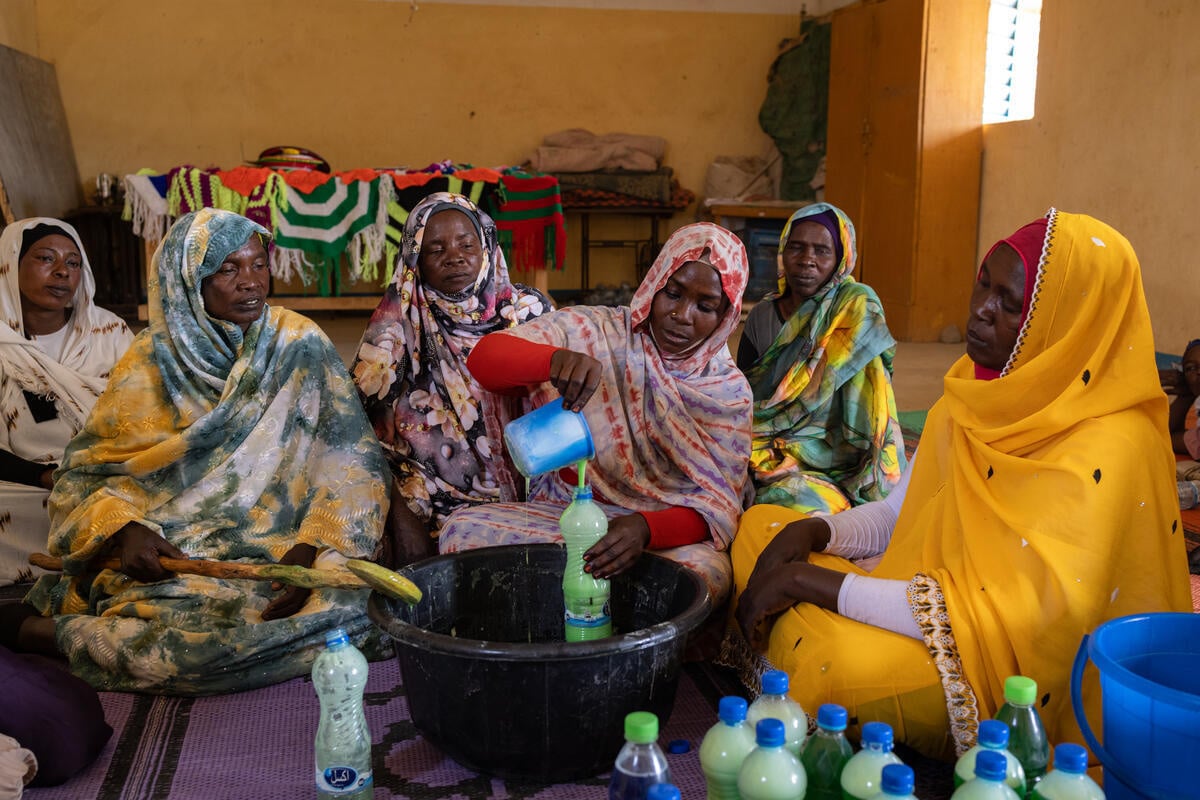 A woman pours liquid soap into a bottle over a large bowl with five other women seated beside her