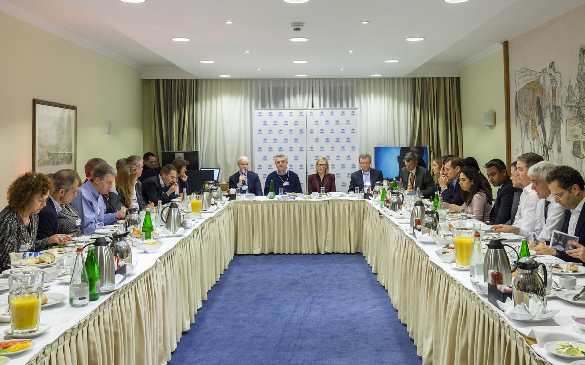 A large group of people seated indoors around a U-shaped table covered in food and drinks