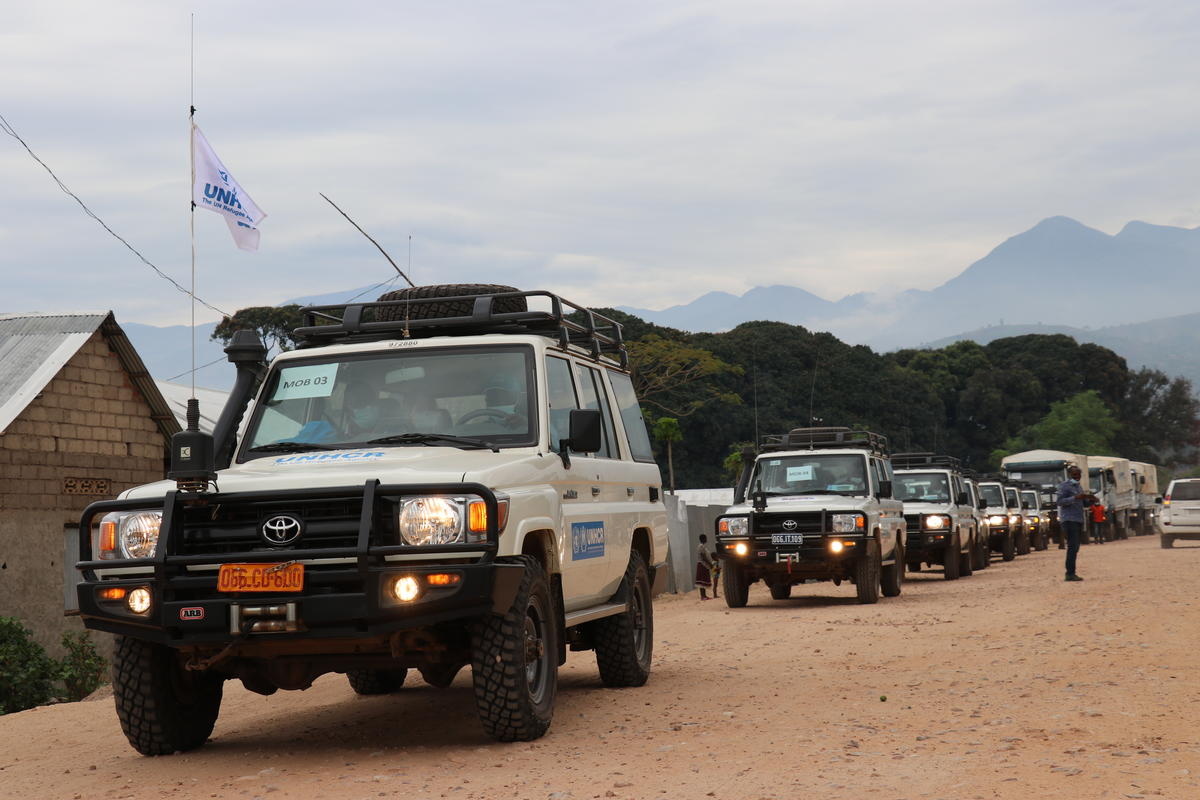 A convoy of UNHCR vehicles on a dirt road with trees and mountains in the background.