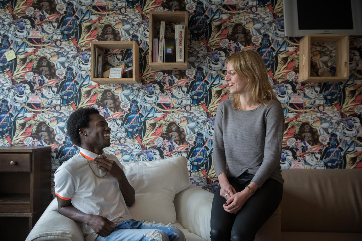 Two friends laughing together on a couch with a colourful wallpaper behind them