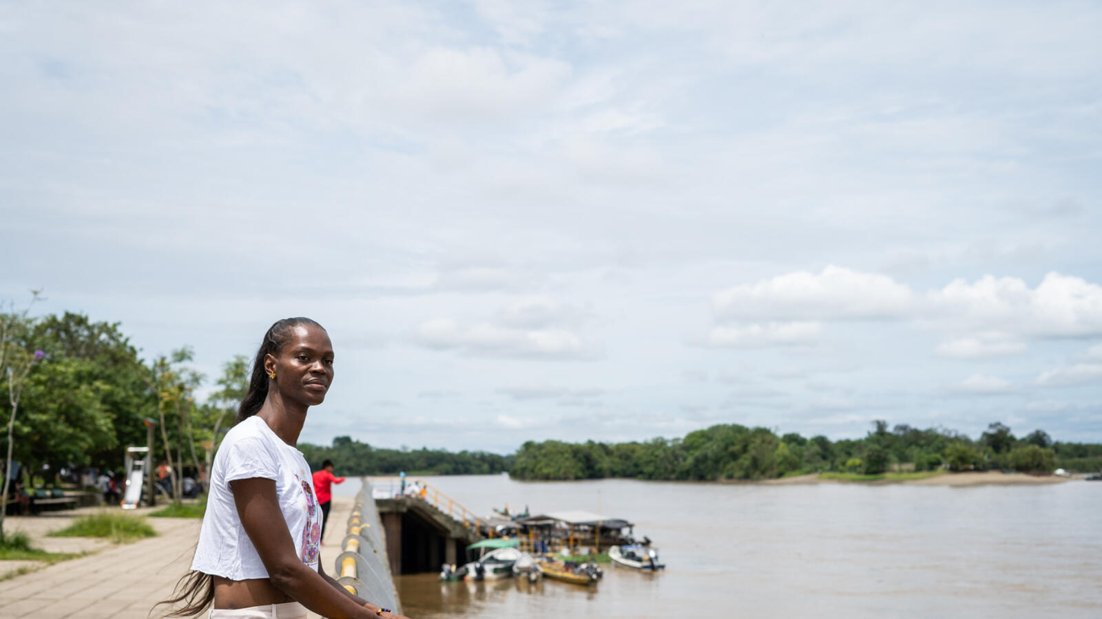 A trans woman leans on a railing looking out over a river.