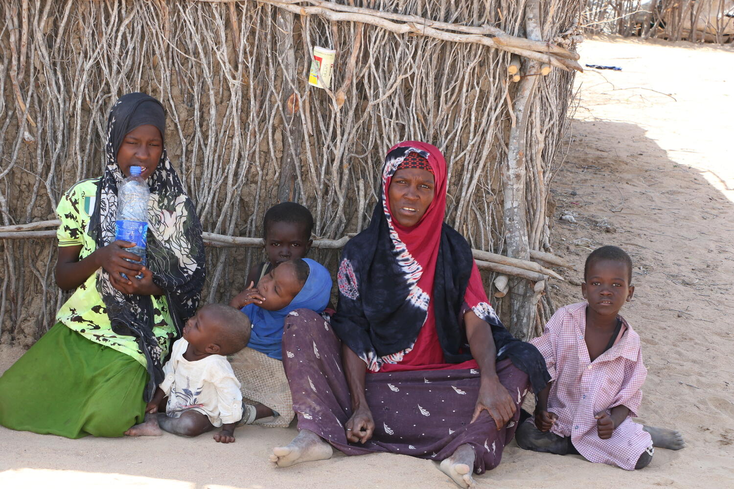 A woman sits on the ground in front of a shelter surrounded by five children.