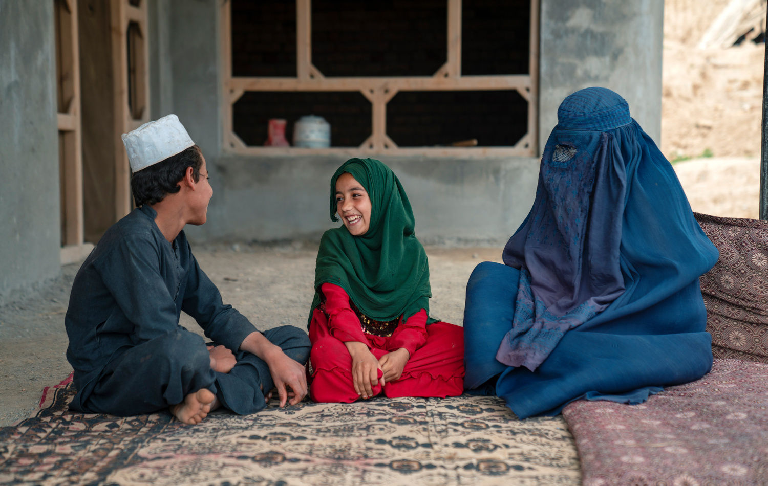 A woman wearing a burka sits on a mat with a girl and a boy,