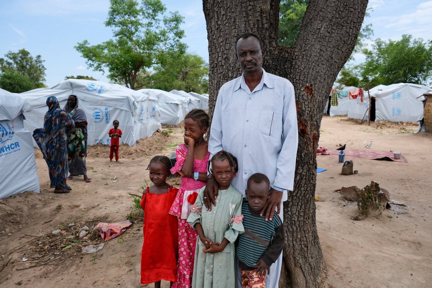 Un homme se tient devant un arbre avec quatre enfants. Des tentes du HCR sont visibles en arrière-plan.