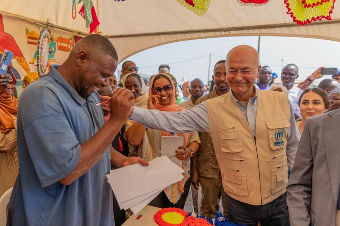 A smiling man drawing a pencil portrait on paper receives a pat on the shoulder from his smiling subject in a tent full of onlookers