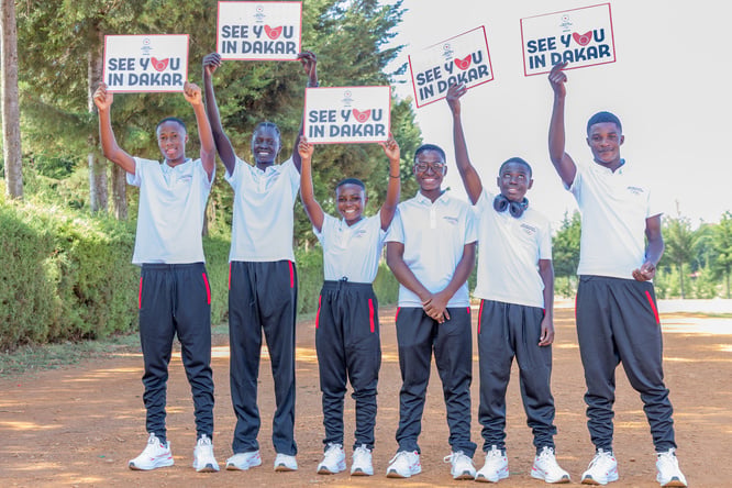 Six jeunes athlètes souriants, vêtus de tenues et de chaussures de sport, brandissent des pancartes sur lesquelles est inscrit « Rendez-vous à Dakar », sur une large route poussiéreuse.