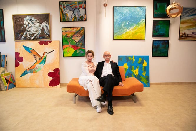 A woman and a man pose on an orange sofa, surrounded by art