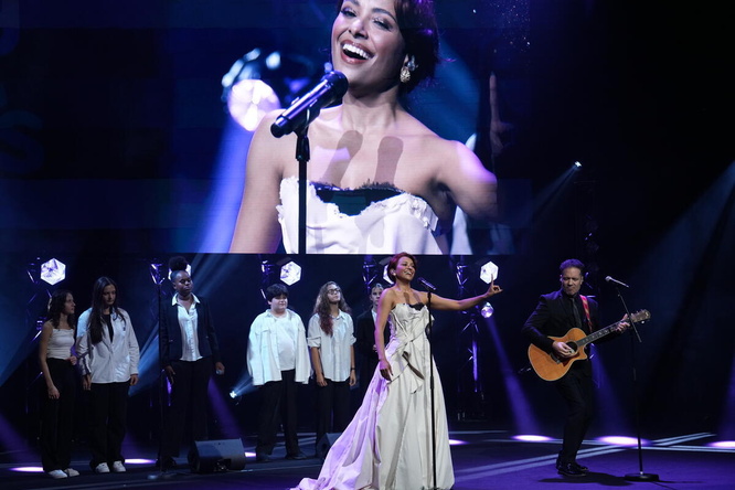 A woman performs onstage with a guitarist and backing singers in front of a giant screen