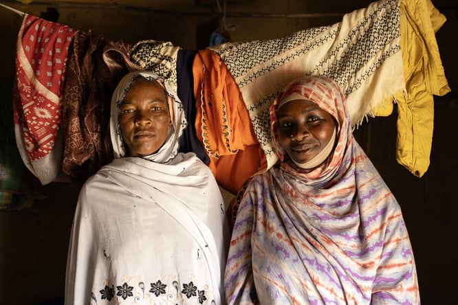Two women stand next to each other inside a room with washing hanging on a line behind them