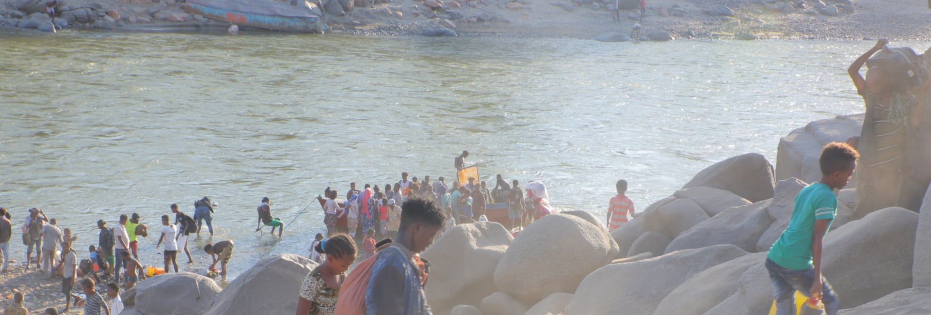 Children carry belongings up a steep, sandy riverbank as people below disembark from a boat
