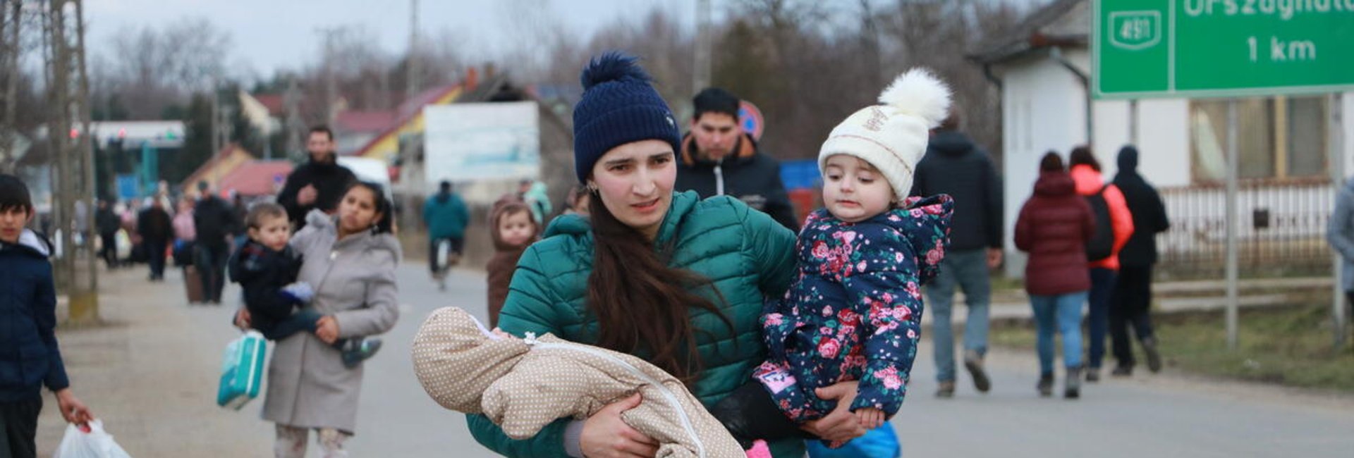 A young woman walks along the side of a road carrying a toddler and a baby in her arms 