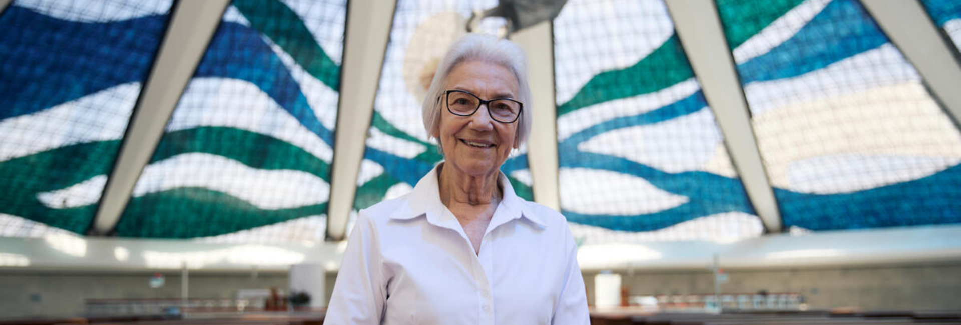 A woman with white hair weaing glasses and a white shirt stands inside a large glass-domed church.