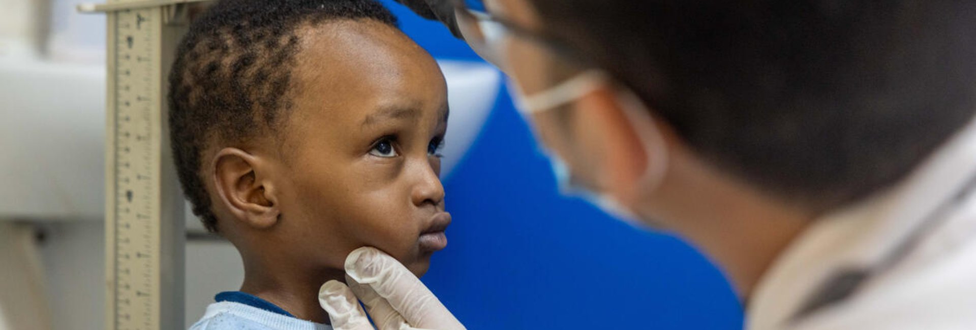 A young boy in a blue sweater stands against a height rule as a man wearing a surgical mask and gloves measures his height.