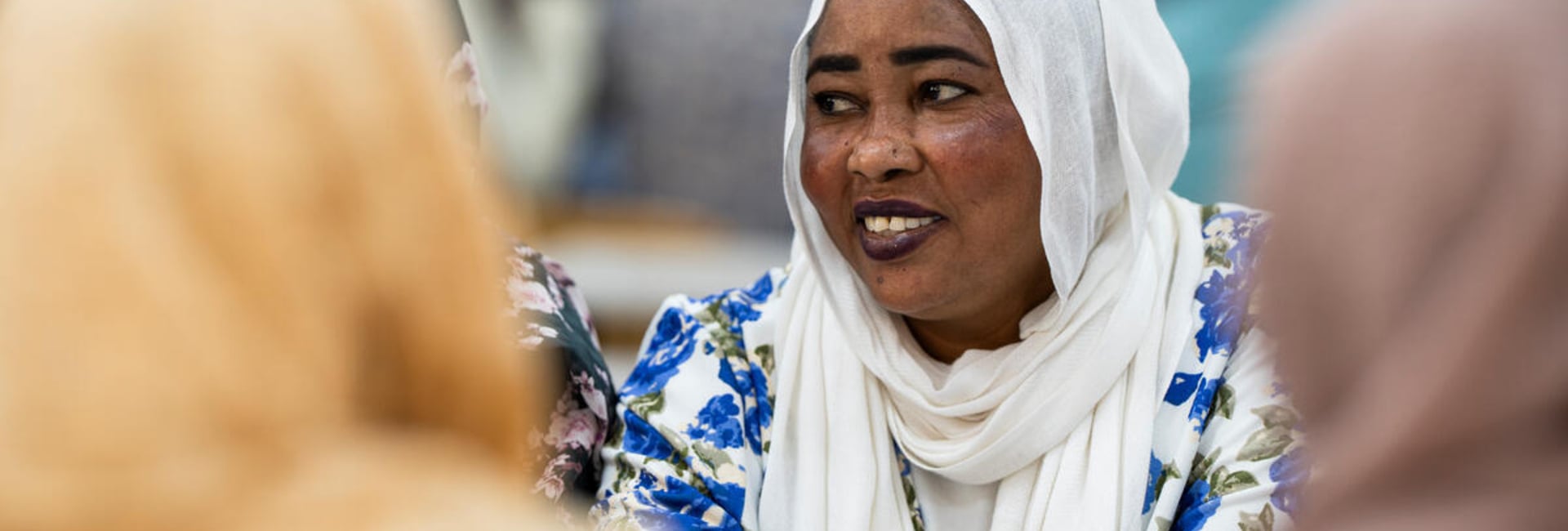 A smiling woman in a white headscarf and blue floral dress sits at a table with other women