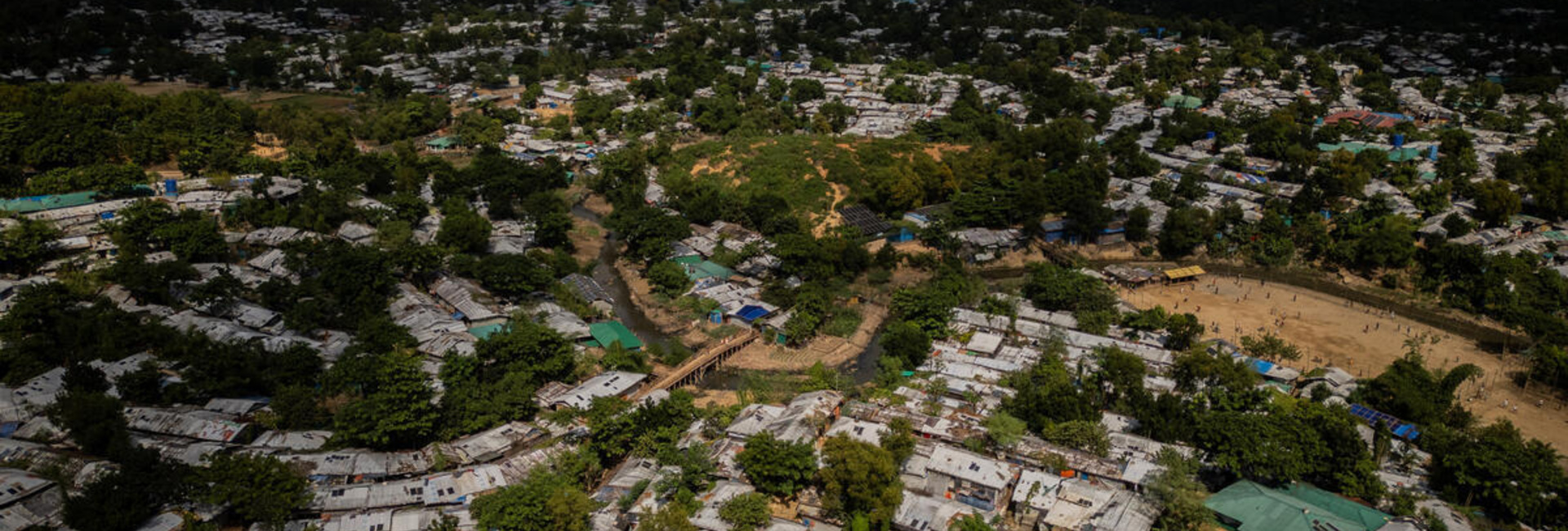 An aerial view of a huge refugee camp, trees throughout, with mountains in the background