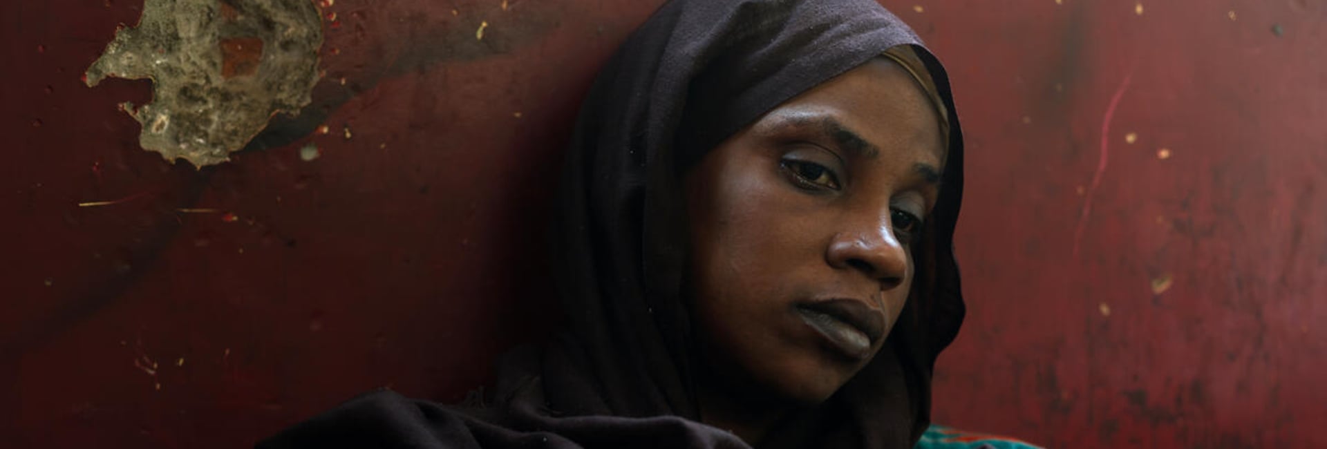 A woman in a colourful dress and a black headscarf sits on the floor leaning against a red-painted, pockmarked wall