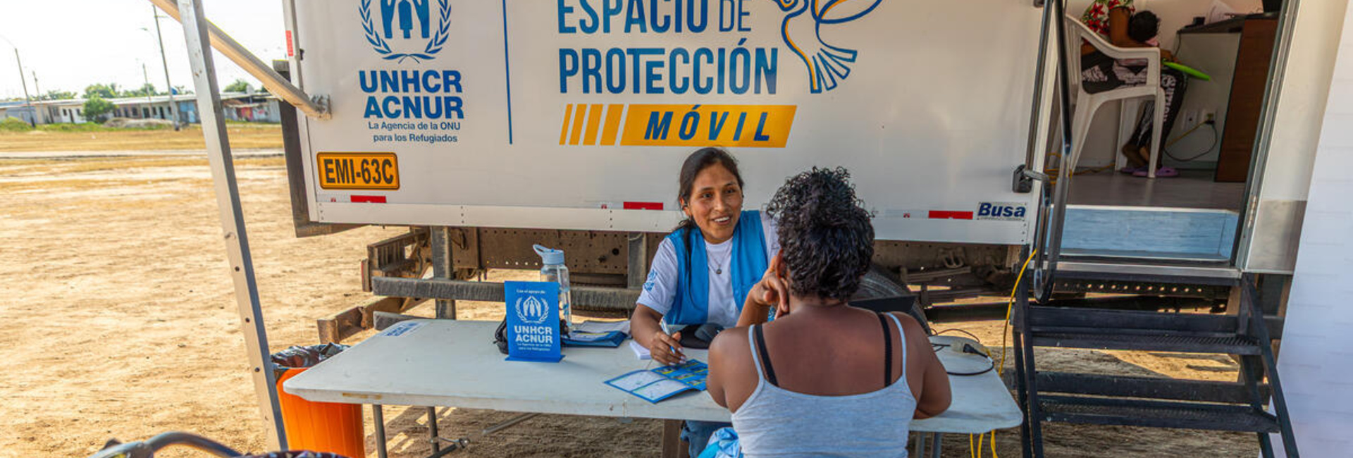 A female member of UNHCR staff speaks to a woman across a table with a baby stroller in the foreground and a truck in the background
