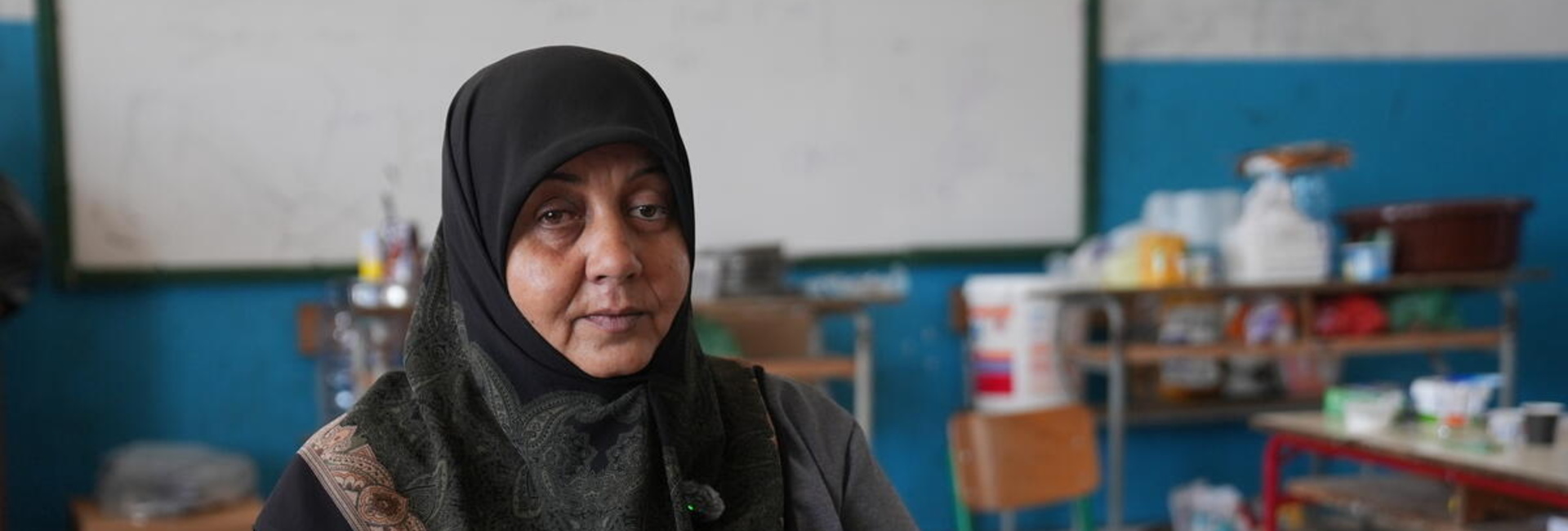 A woman in a headscarf sits in a school classroom with household items on desks in the background