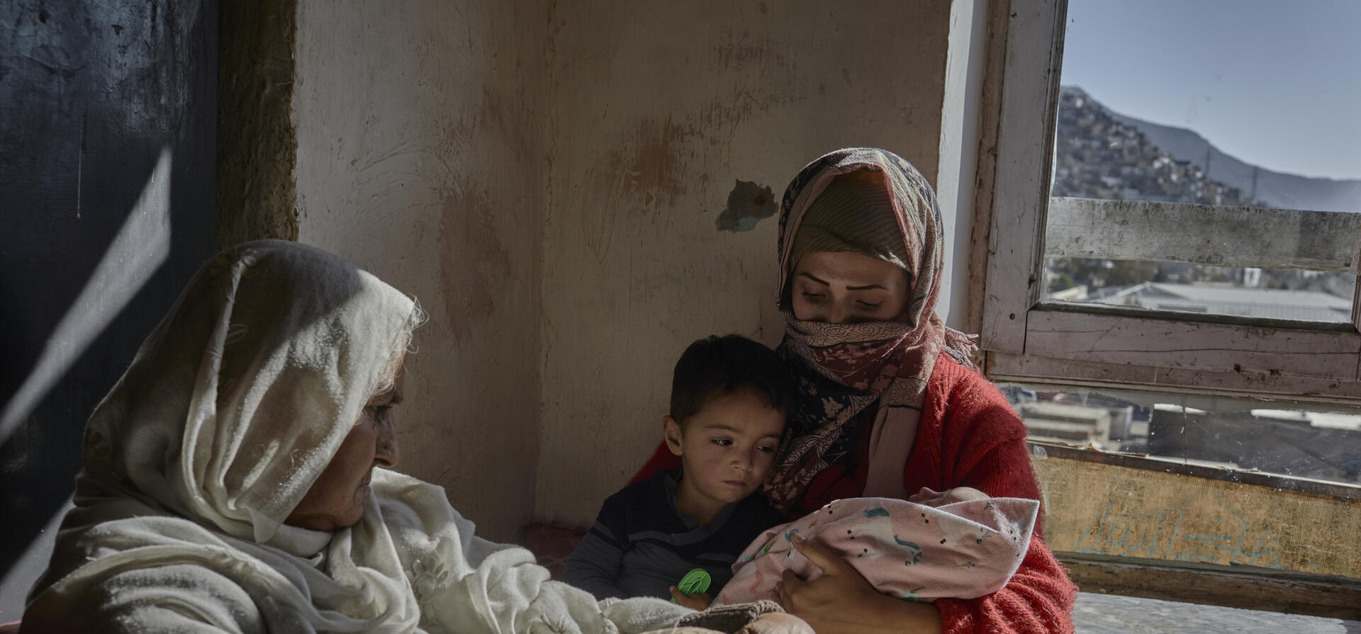 A mother cradles her baby in a bare room with her son and mother looking on
