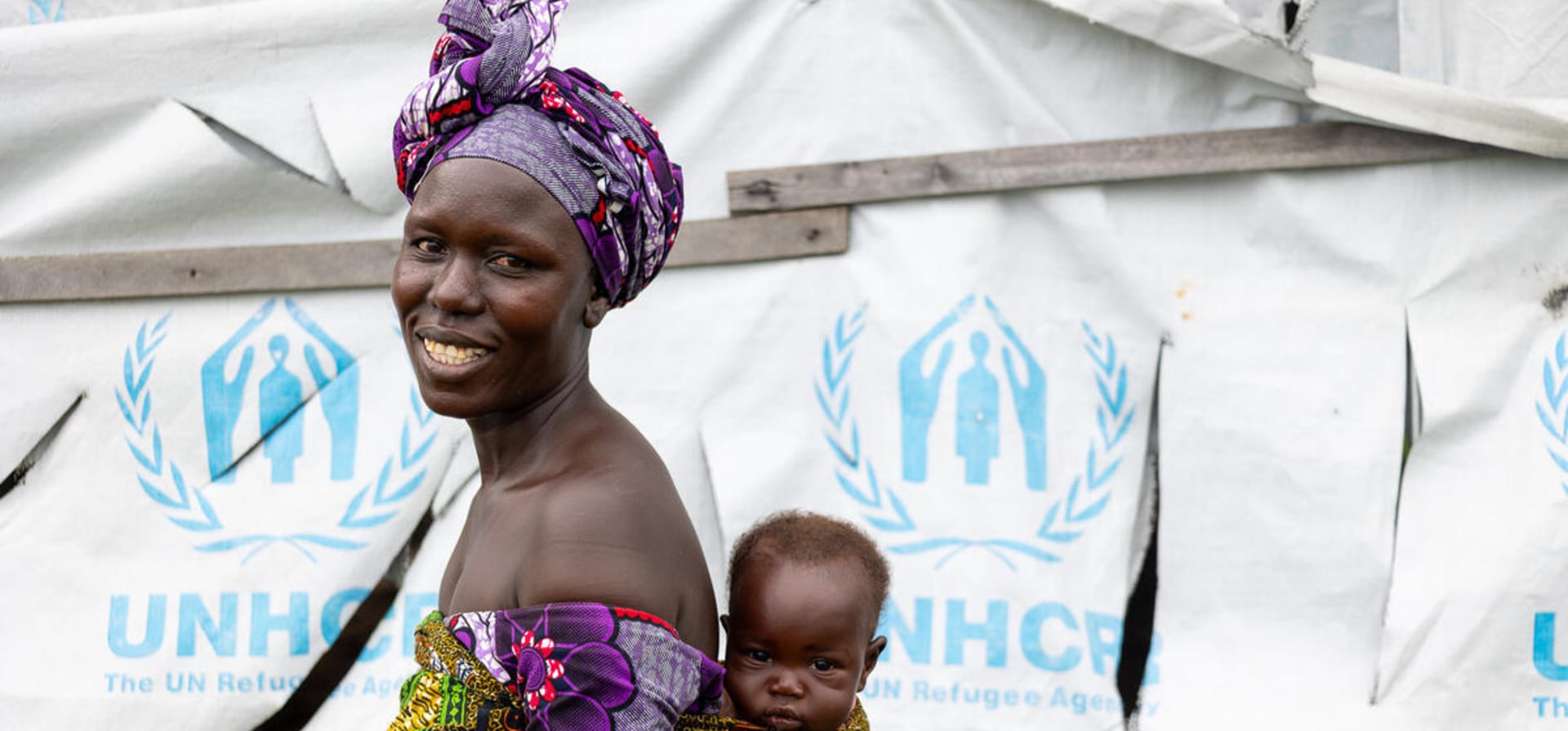 A smiling woman carries her baby boy on her back in front of a UNHCR tent.
