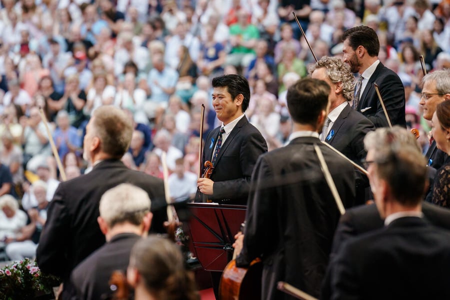 Musicians part of an orchestra stand up holding their instruments, in front of a crowd.
