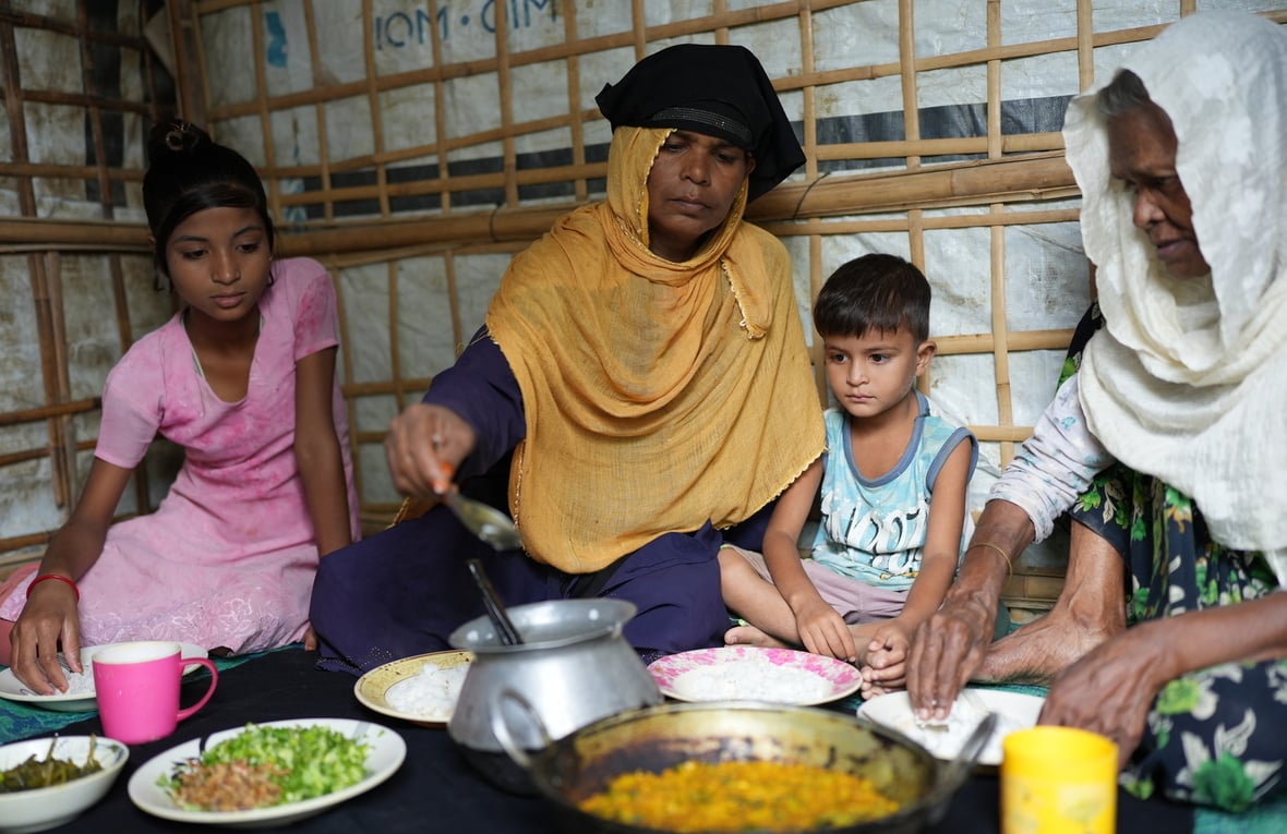 A mother and a grandmother eat a meal sitting on the floor of their shelter with two young children.