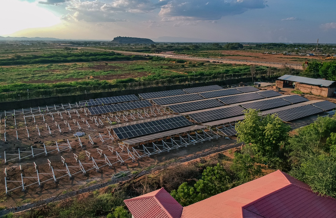 An aerial view of a partially constructed solar farm with a red-roofed building in the foreground and a savannah landscape stretching to the horizon.  