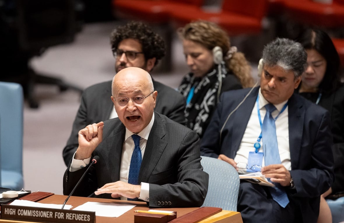 A man gestures while speaking behind a desk with people sitting in the background