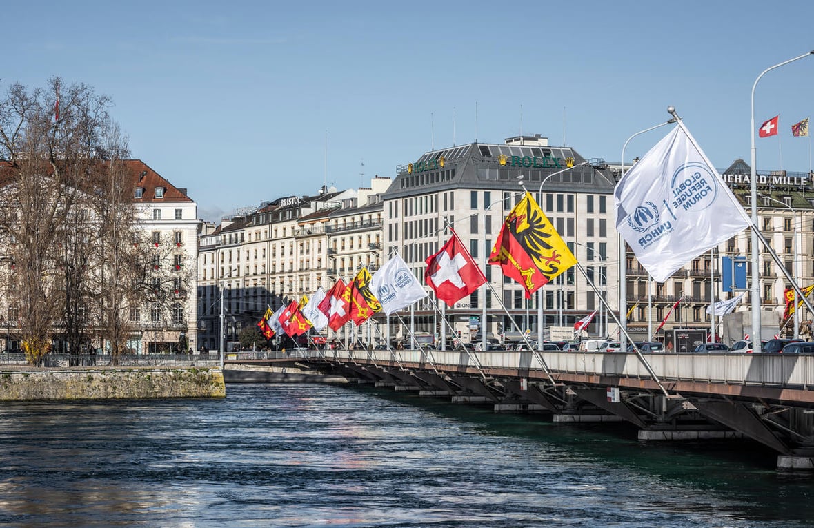 Flags fly along the length of a bridge over a wide river with buildings in the background