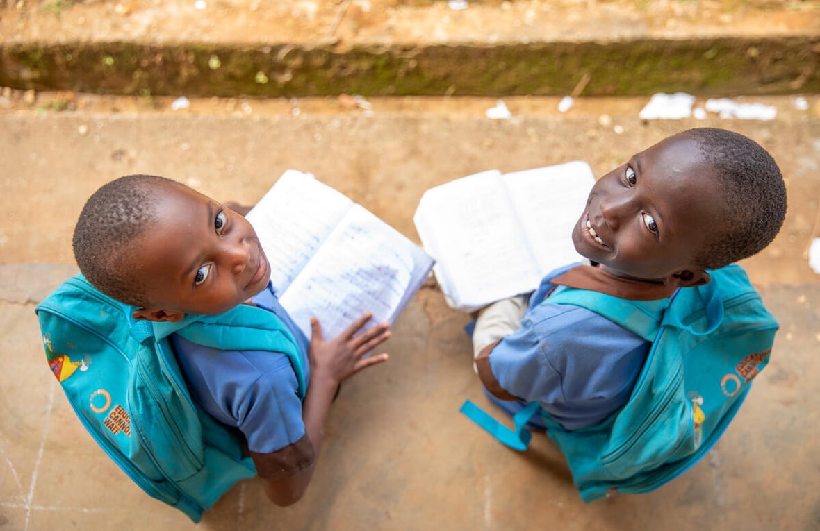 Two girls, 7- and 8-years-old, from Central African Republic smile up at the camera from where they're sitting outside, wearing matching 'Education cannot wait' backpacks and holding open books.