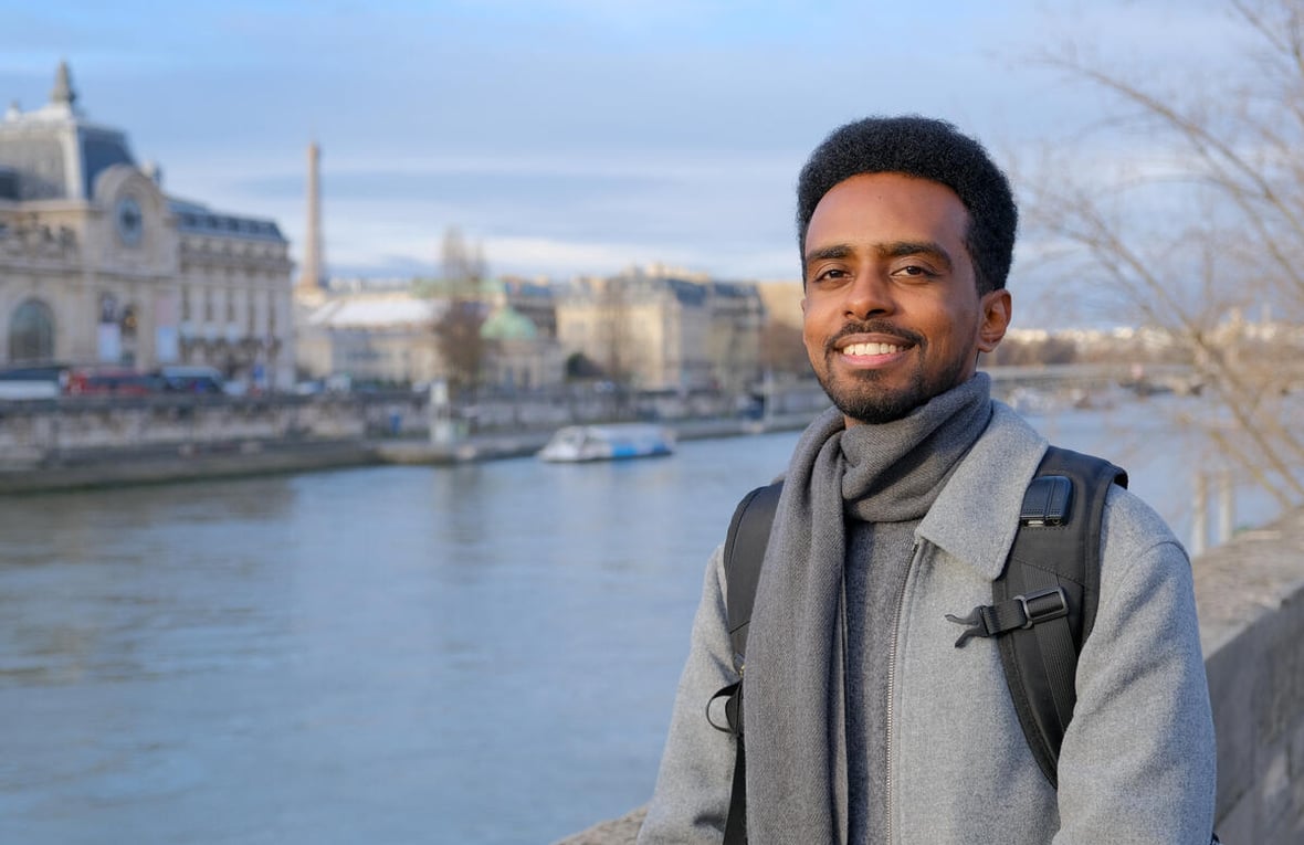 A man wearing a grey overcoat and backpack stands on a riverside with historic buildings on the opposite bank 