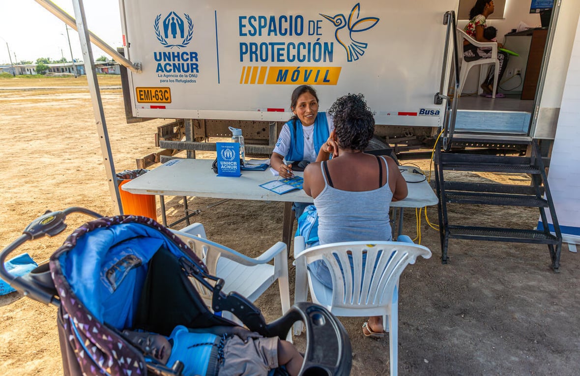 A female member of UNHCR staff speaks to a woman across a table with a baby stroller in the foreground and a truck in the background