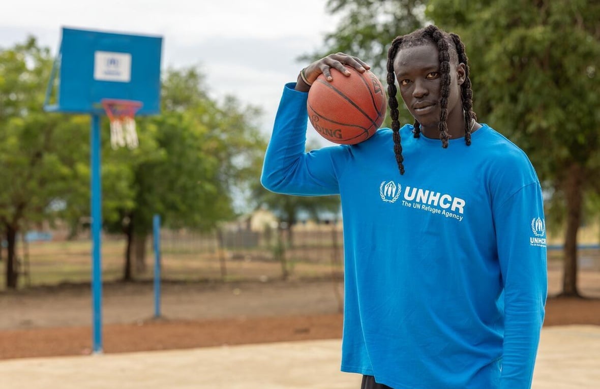 A South Sudanese man holds a basketball on his shoulder, a basketball court behind him