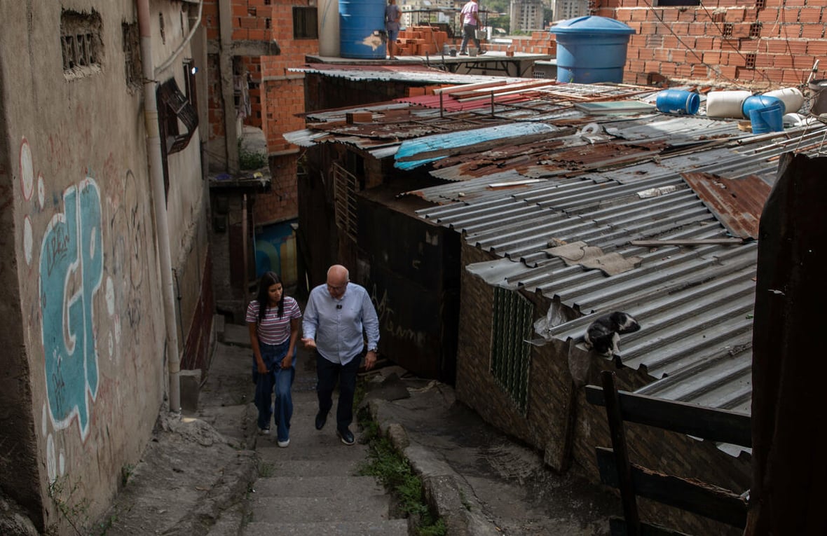 A man and a woman walk side by side up the staircase of a narrow street beside tin-roofed buildings 