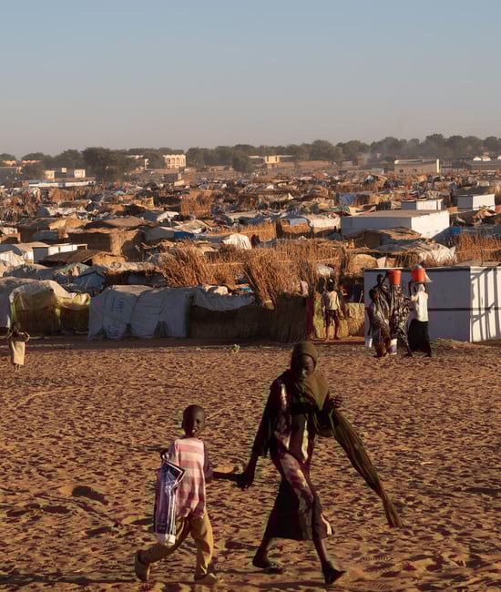 A woman holds a child's hand as they walk past a refugee tent settlement in a dusty field.