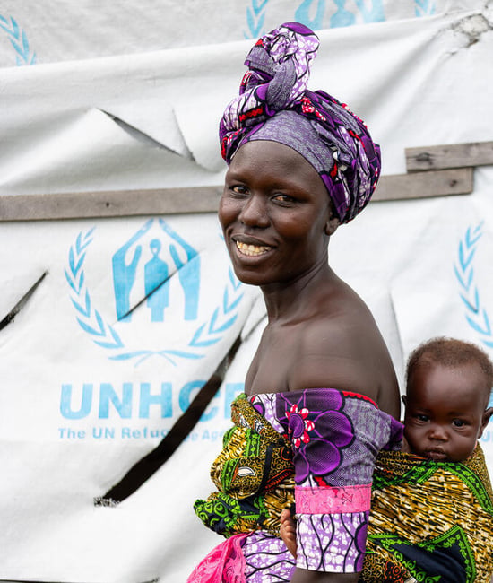 A smiling woman carries her baby boy on her back in front of a UNHCR tent.