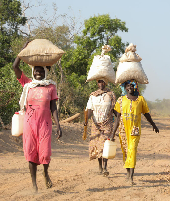 Three friends, Sudanese refugee women, walk together along a dirt road, bags balanced on their heads, smiling and laughing.