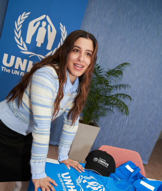 Marina pictured in front of a UNHCR banner, standing by a table with UNHCR t-shirts, caps and books.