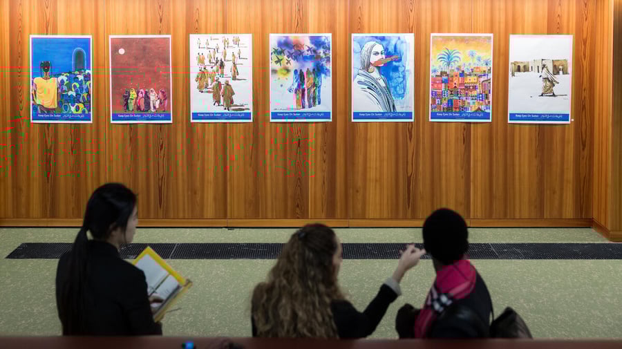 Three persons sit on a couch, looking at several artworks displayed on a wall in front of them.