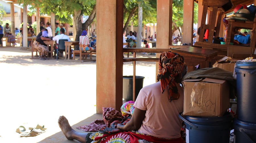 A woman sits in the shade on the wooden terrace of a school, leaning back against boxes of aid supplies 