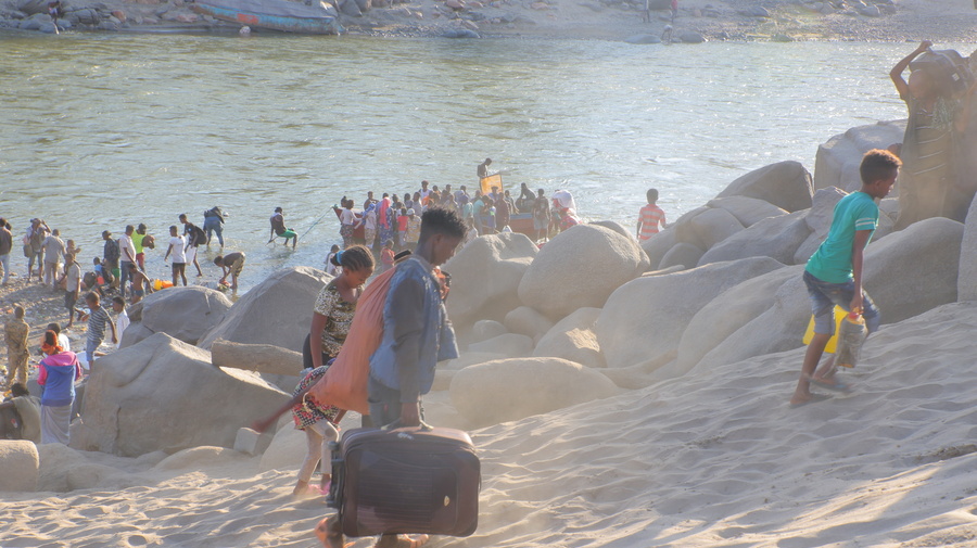 Children carry belongings up a steep, sandy riverbank as people below disembark from a boat